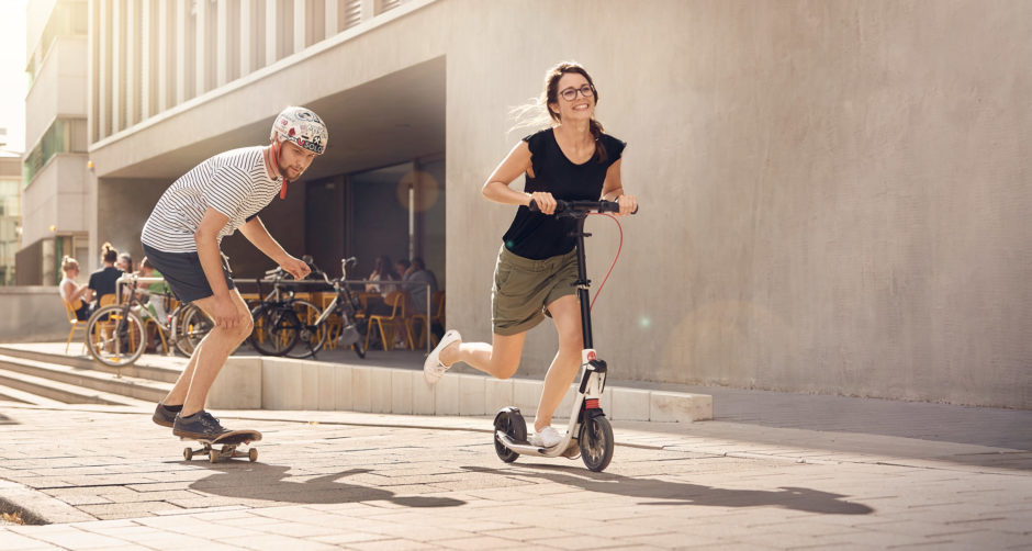 Mann auf Skateboard und Frau auf Tretroller unterwegs auf Fußweg
