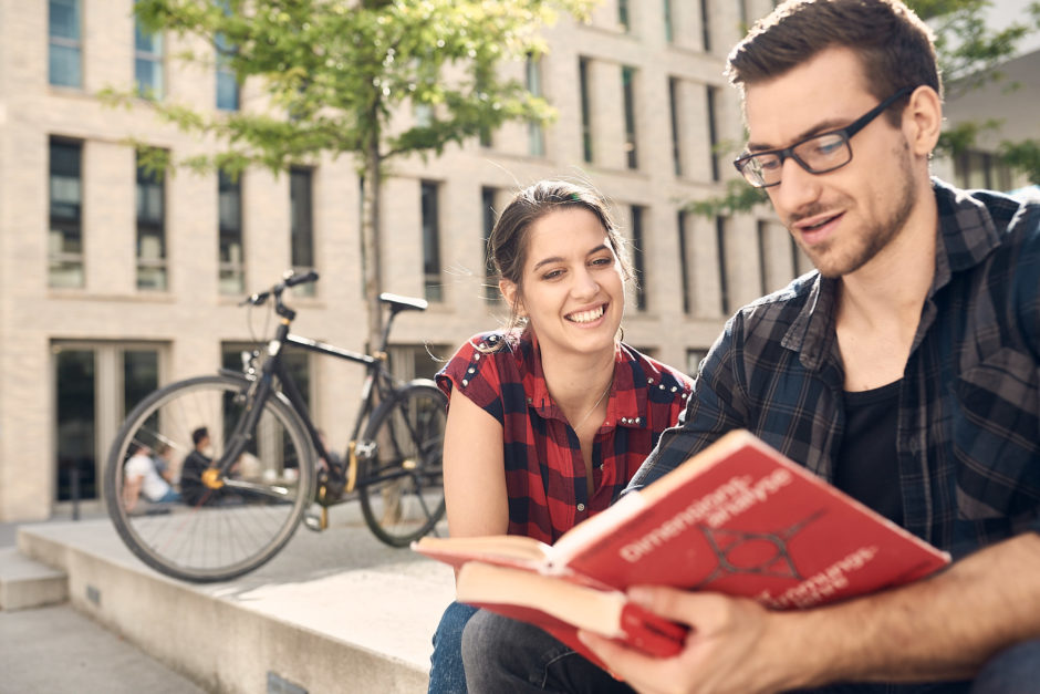 Studierende am Campus lesen in Fachbuch, im Hintergrund abgestelltes Fahrrad